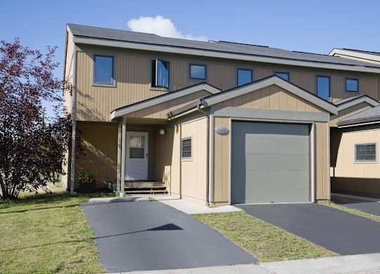 A well-kept Birchwood townhome exterior featuring a tan two-story façade with gray trim, a single-car garage, and a small covered entryway with a white front door. The freshly paved driveway and green lawn frame the home, while a tree and bright summer skies add curb appeal and a welcoming neighborhood feel.