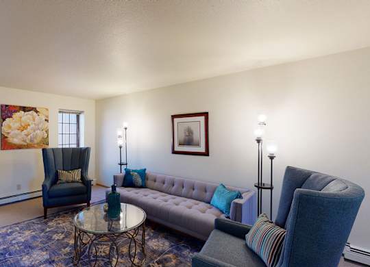 A bright living room at Birchwood Apartments with a tufted sofa, two upholstered high-back chairs, and a round glass coffee table on a patterned area rug. Wall art, accent pillows, and floor lamps add color and warmth, while two windows bring in natural light from outside.