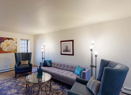 A bright living room at Birchwood Apartments with a tufted sofa, two upholstered high-back chairs, and a round glass coffee table on a patterned area rug. Wall art, accent pillows, and floor lamps add color and warmth, while two windows bring in natural light from outside.