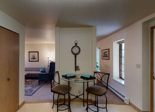 A cozy dining nook at Birchwood Apartments featuring a round glass table with two cushioned chairs set against a dividing wall decorated with a vintage key-shaped wall clock, with views into the adjoining living room and hallway lined with windows.