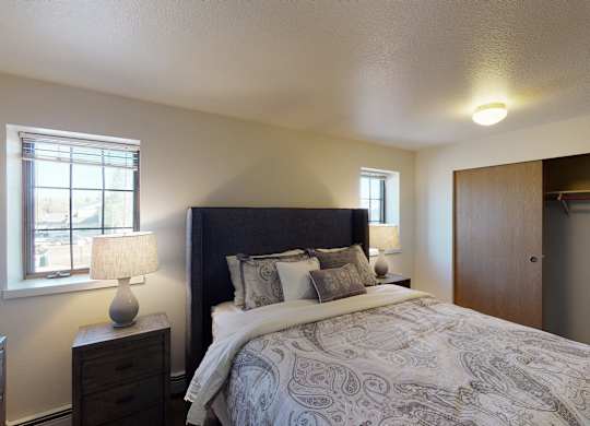 A bright Birchwood bedroom featuring a dark upholstered headboard with paisley bedding and layered pillows centered against a neutral wall. Two matching lamps on wooden nightstands sit beside the bed beneath twin windows. The room includes soft carpet and a wide closet with sliding wooden doors partially open.