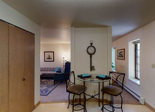 A cozy dining nook at Birchwood Apartments featuring a round glass table with two cushioned chairs set against a dividing wall decorated with a vintage key-shaped wall clock, with views into the adjoining living room and hallway lined with windows.