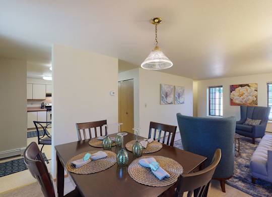 Dining area at Birchwood Apartments featuring a dark wood table set for four with woven placemats and folded napkins, opening into a cozy living room with upholstered seating, floral artwork, and large windows letting in natural light.