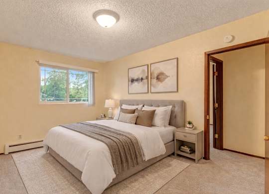 A serene Brighton Woods bedroom featuring a large window that frames green treetops and fills the room with daylight. A plush bed dressed in white linens and warm earth-tone pillows sits against a soft yellow wall, complemented by matching nightstands, soft carpet, and simple abstract artwork for a peaceful retreat.