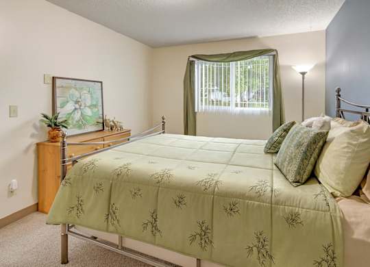 A charming Brighton Woods bedroom decorated in soft green tones with a neatly made bed featuring botanical-patterned bedding and layered pillows. A large window framed by matching curtains brings in natural light, while a light wood dresser with artwork and a tall floor lamp add warmth to the cozy space.