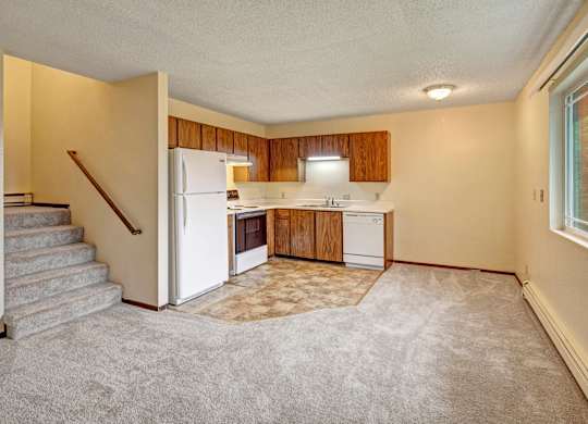 A bright Brighton Woods kitchen and dining space featuring warm wood cabinets, white appliances, and a neutral countertop arranged in an L-shape. Soft carpeting flows into the room from an adjacent open living area, with a cozy staircase leading upstairs and a large window bringing in natural light for an inviting everyday layout.