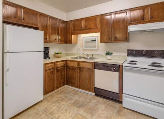 A warm kitchen here at Castle featuring rich wood cabinetry, a white refrigerator and stove, and a spacious L-shaped countertop with a double sink. Simple décor like a framed print, a coffee maker, and bowls of fruit adds inviting touches, while soft lighting brightens the cozy layout.