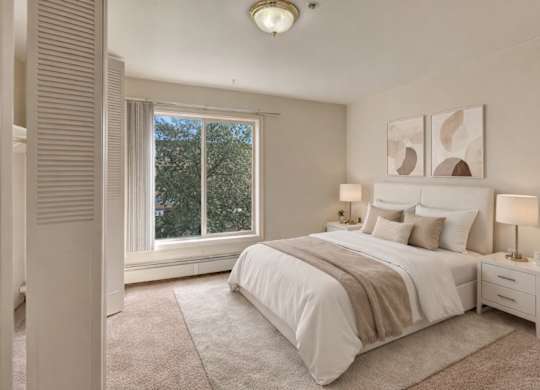 A serene bedroom here at Castle showcases a neatly made bed with white bedding, soft tan throws, and coordinating pillows set against two modern abstract prints. Plush carpet runs wall to wall while large windows bring in natural light and showcase leafy treetop views beside warm bedside lighting on matching nightstands.