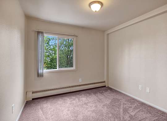 A cozy bedroom here at Castle features soft neutral walls and plush carpeting that creates a warm and inviting feel. A large window brings in natural light and frames lush green treetops outside, while simple baseboard heating and an uncluttered layout offer a versatile space ready for personal furniture and décor.