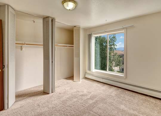 A spacious bedroom here at Castle features soft carpet flooring and warm neutral walls with a large window drawing in sunlight and views of leafy trees and nearby rooftops. Wide double closets with folding doors offer generous storage, while the open layout provides plenty of room for furniture and personal touches.