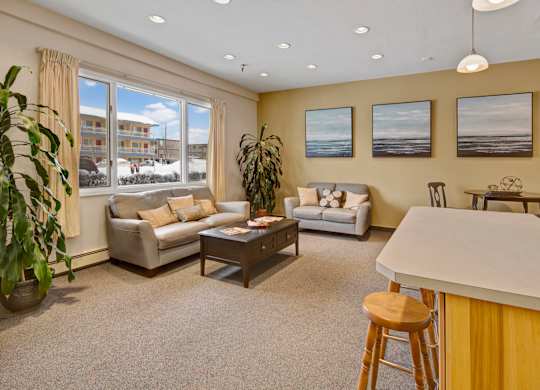 A bright community lounge here at Century Plaza featuring two cozy loveseats with soft throw pillows, a dark wood coffee table, and large windows bringing in natural light. Three coastal art pieces line the yellow accent wall, with plants, bar seating, and a small dining table completing the inviting space.