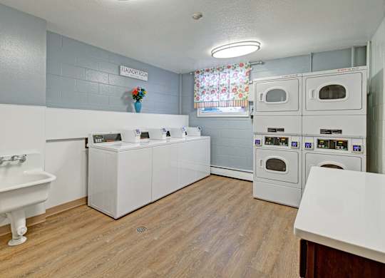A bright on-site laundry room here at Century Plaza featuring multiple top-loading washers, stacked dryers, a deep utility sink, wood-look flooring, and a window with colorful curtains for natural light.