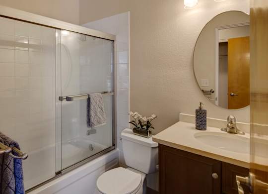A modern bathroom here at Century Plaza featuring a clean tiled shower with sliding glass doors, a white toilet, and a spacious vanity with a round mirror and brushed-nickel fixtures. Neutral walls, soft lighting, and small décor accents create a bright and polished space.