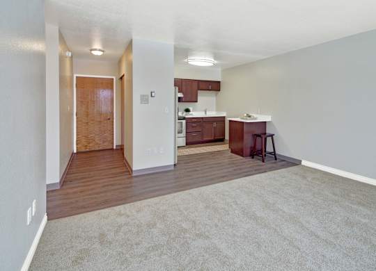 A spacious living area here at Century Plaza featuring a blend of soft carpet and rich wood-style flooring that transitions into the kitchen. The open layout includes a breakfast bar with stool seating, warm wood cabinetry, and modern lighting, while the front entry hallway adds convenience and flow to the space.
