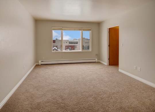 A bright living space here at Century Plaza showcasing an open layout with neutral wall tones and plush carpeting. A large picture window lets in natural light and offers a view of nearby buildings under a clear blue sky, while an adjoining doorway connects the room to the rest of the apartment.