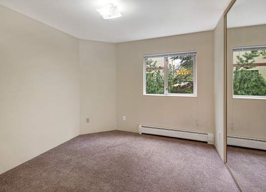 Cozy empty bedroom here at Chugach South with beige walls, carpeted floors, a window looking out to trees, and mirrored closet doors.