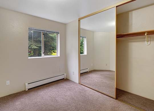 A bright empty bedroom here at Chugach South with beige walls, carpet flooring, large mirrored closet doors, and a window looking out to trees.