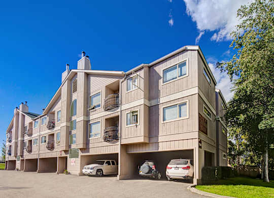 A sunny exterior view of Chugach South showing a three-story tan apartment building with small balconies, carports with parked vehicles, and trees and blue sky in the background.