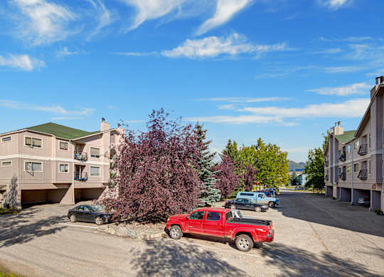 A sunny exterior view of Chugach South showing tan multi-story apartment buildings with covered parking, a red pickup truck and other vehicles in the lot, purple leafy trees, and mountains visible in the distant background under a bright blue sky with wispy clouds.