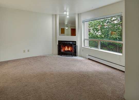 A cozy living room here at Chugach South featuring light walls, beige carpeting, a lit wood-burning fireplace with a mirror above it, a large window overlooking green trees, and a fire extinguisher mounted on the wall.