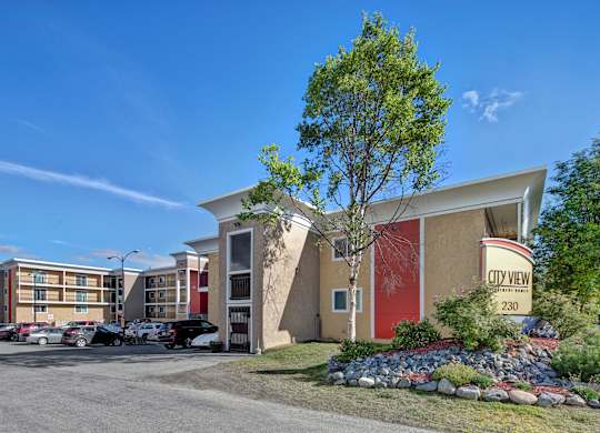 A sunny exterior view of City View Apartment Homes showcasing modern multi-story buildings with bold red and tan accents, balconies, and well-kept landscaping framed by rocks, shrubs, and trees. A clear blue sky, parked cars, and the welcoming entrance sign create a bright and inviting first impression of the community.