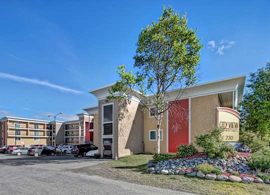A sunny exterior view of City View Apartment Homes showcasing modern multi-story buildings with bold red and tan accents, balconies, and well-kept landscaping framed by rocks, shrubs, and trees. A clear blue sky, parked cars, and the welcoming entrance sign create a bright and inviting first impression of the community.