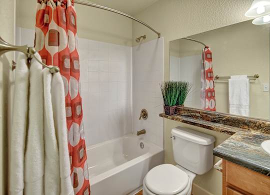 A bright bathroom here at City View featuring a white tub and shower with a curved rod and bold orange patterned curtain, a wide granite countertop with natural earth tones, and wood cabinetry below. A large mirror stretches across the wall, complemented by soft lighting, fluffy white towels, and a small faux plant for a touch of color.