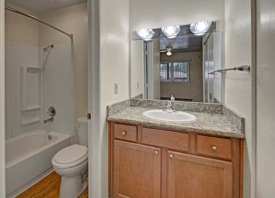 A clean and functional bathroom here at Club at Eagle Pointe featuring a granite-style vanity countertop with a single sink, warm wood cabinetry, and a large wall mirror with bright overhead lighting. The space includes a tub and shower combination with built-in shelving, durable flooring, and a layout designed for everyday comfort and convenience.