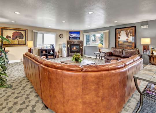 A spacious, inviting community lounge here at Club at Eagle Pointe featuring plush leather sectional seating, a cozy fireplace with a mounted TV, warm accent lighting, large windows for natural light, and tasteful artwork throughout, creating a comfortable gathering space for relaxing, socializing, or enjoying quiet time indoors.