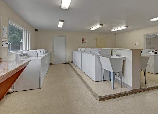 A clean, well-lit on-site laundry room here at Club at Eagle Pointe featuring multiple washers and dryers, a utility sink, wide walkways, bright overhead lighting, and durable flooring, providing residents with a convenient, functional space for everyday laundry needs.