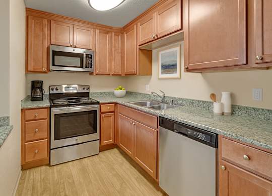 A warm, functional kitchen here at Club at Eagle Pointe featuring natural wood cabinetry, granite-style countertops, and stainless steel appliances including an oven, microwave, and dishwasher. Durable wood-style flooring and ample counter space create a practical layout that supports everyday cooking and comfortable meal preparation.