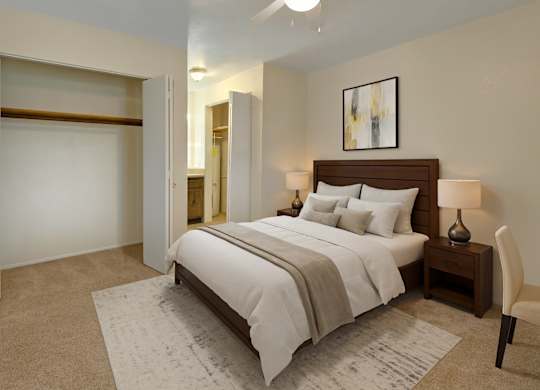 A spacious, inviting bedroom here at Club at Eagle Pointe featuring soft neutral walls, plush carpet flooring, and a comfortable bed with layered white bedding and accent pillows. An open closet with ample hanging space, bedside tables with lamps, and an adjacent bathroom create a functional, relaxing retreat designed for everyday comfort.