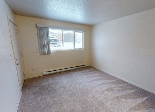 A spacious, sunlit bedroom here at Club at Eagle Pointe featuring soft neutral walls, plush carpet flooring, and a wide window with vertical blinds that bring in natural light. A baseboard heater runs beneath the window, and the simple rectangular layout offers ample space for a bed, dresser, and additional furnishings.