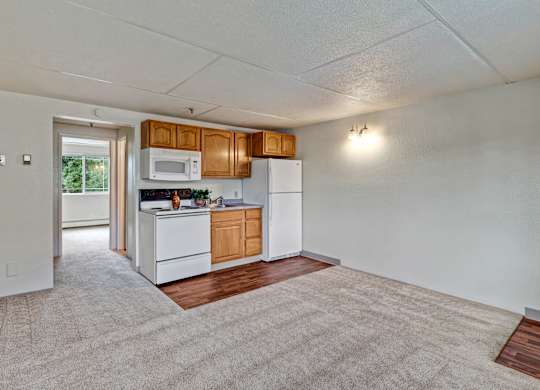 A bright open living area here at College View featuring plush carpet flooring, neutral wall colors, and an efficient kitchen with warm wood cabinetry, white appliances, and easy-care vinyl flooring. The open layout provides flexibility for living and dining furniture, smooth flow between spaces, and a comfortable setting for everyday living.