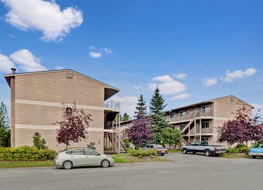 A welcoming exterior here at College View showcasing well-maintained apartment buildings with neutral siding, exterior staircases, and private balconies. Mature trees, landscaped greenery, and open parking areas create an inviting community setting, while clear walkways and open skies highlight the comfortable residential atmosphere.