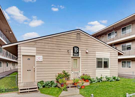 A charming exterior view here at College View featuring a well-kept community building with neutral siding, clearly marked entrances, and surrounding apartment structures with balconies. Lush green lawns, colorful potted plants, and tidy walkways create a welcoming residential atmosphere under bright blue skies.