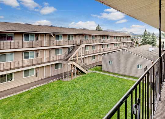 A bright courtyard view here at College View showcasing multi-story apartment buildings with exterior walkways, railings, and staircases overlooking a well-maintained green lawn. Neutral siding, large windows, and open corridors create a clean, welcoming community layout with mountain views and blue skies visible in the background.