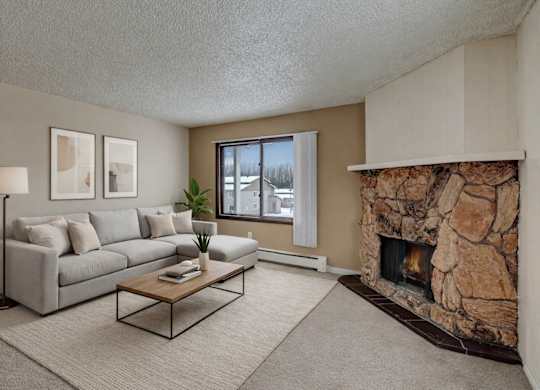 A cozy living room here at Conifer Grove featuring a light gray sectional sofa with neutral pillows, a simple wooden coffee table, and a soft area rug that defines the seating space. A large window brings in natural light, while a striking stone fireplace adds warmth and character to the room.