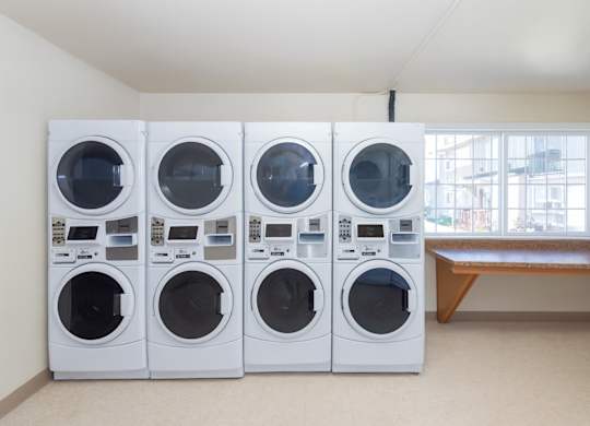 A bright community laundry room here at Conifer Grove featuring stacked front-loading washers and dryers, a large window providing natural light, and a convenient folding counter. The space is clean, open, and designed to make laundry tasks simple and efficient for resident