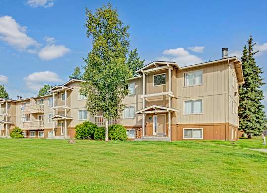 A sunny exterior view here at Conifer Grove showcasing a multi-story apartment building with neutral siding, private balconies, and large windows, surrounded by well-maintained green lawns, mature trees, and a bright blue sky that highlights the community’s peaceful residential setting.