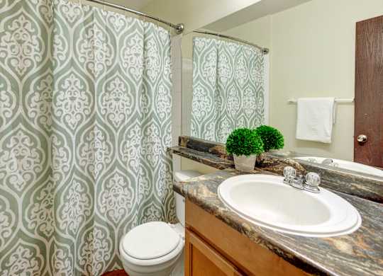 A fresh bathroom here at Conifer Grove featuring a clean tub and shower combination with a patterned shower curtain, a spacious vanity with wood cabinetry, and a durable countertop with an integrated sink. The space includes a large wall mirror, neutral wall tones, and simple fixtures that create a functional and comfortable daily routine.