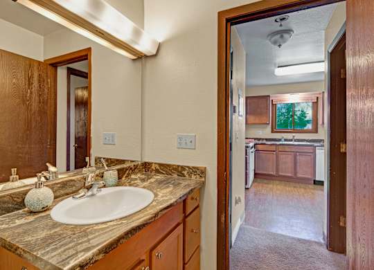 Bright bathroom here at Conifer Grove featuring a spacious vanity with wood cabinetry, a durable stone-style countertop, and an integrated sink beneath a large wall mirror. The layout offers ample counter space, warm neutral walls, and convenient access to the kitchen area, creating a functional and comfortable daily routine.