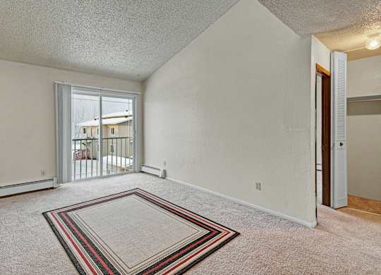 Bright living room here at Conifer Grove featuring soft carpeting, neutral walls, and a large sliding glass door that opens to a private balcony and allows in natural light. The open layout offers flexible space for seating or a dining area, with easy access to nearby rooms and a comfortable, inviting feel.