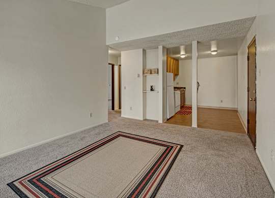 Spacious living room here at Conifer Grove featuring soft carpet flooring, neutral walls, and an open layout that flows into the kitchen and dining area. The room includes built-in shelving, overhead lighting, and a defined entry space, offering flexible options for seating and everyday living.