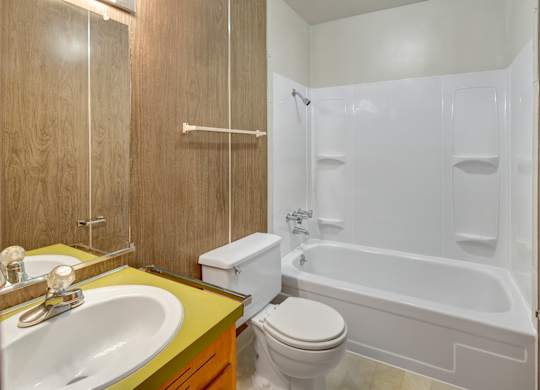 A clean bathroom here at Country Lane featuring a white tub and shower surround with built-in shelving, a single-sink vanity with a yellow countertop, a mirrored medicine cabinet, wood-paneled walls, and durable flooring designed for easy everyday use.