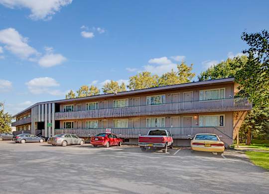 A bright exterior view here at Country Lane showcasing a two-story apartment building with covered walkways, private entry doors, large windows, on-site parking, and mature trees surrounding the community under a clear blue sky.