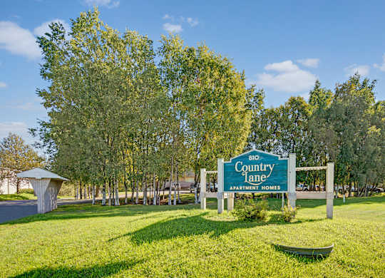 A welcoming community sign here at Country Lane displaying the Country Lane Apartment Homes name and address, set against a landscaped green lawn with mature trees and a clear blue sky, marking the entrance to the apartment community.
