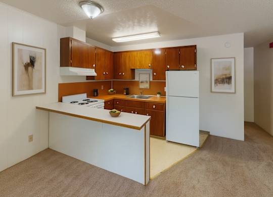 A functional kitchen here at Country Lane featuring warm wood cabinetry, white appliances, and an open breakfast bar overlooking the living area, with neutral flooring, simple wall art, and bright overhead lighting creating a clean and practical apartment layout.