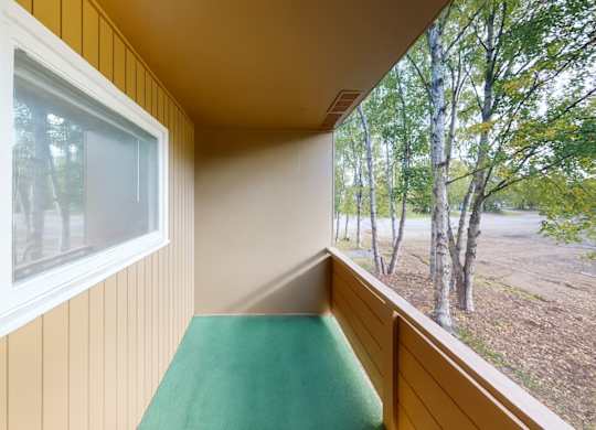 A covered balcony here at Country Lane offering a private outdoor space with neutral siding, sturdy railing, and a soft green floor surface, providing a peaceful spot to enjoy fresh air and views of surrounding trees while extending the apartment’s living space.