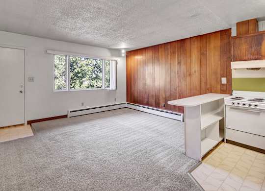 A bright living room here at Country Lane featuring soft carpet flooring, a large window that brings in natural light, and a distinctive wood-paneled accent wall, with an open connection to the kitchen peninsula that creates a functional and welcoming open-concept living space.